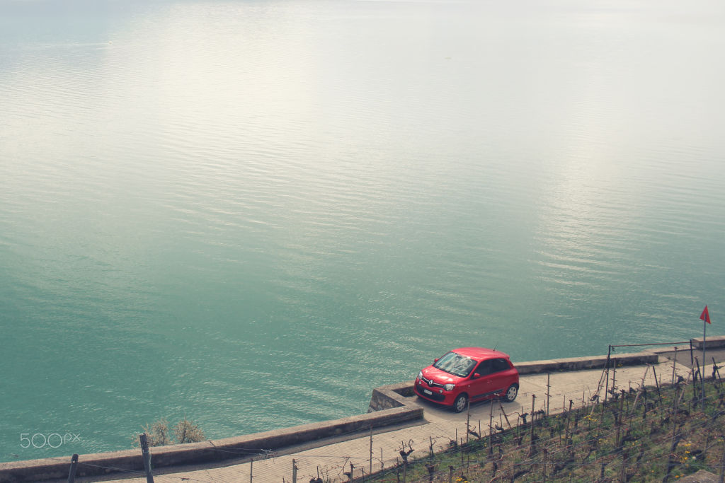 A red car on a lakeside road above turquoise water