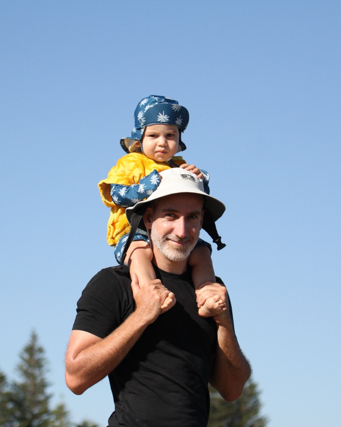 Pierre Sauvignon under a blue sky, his son on his shoulders, both in sun hats