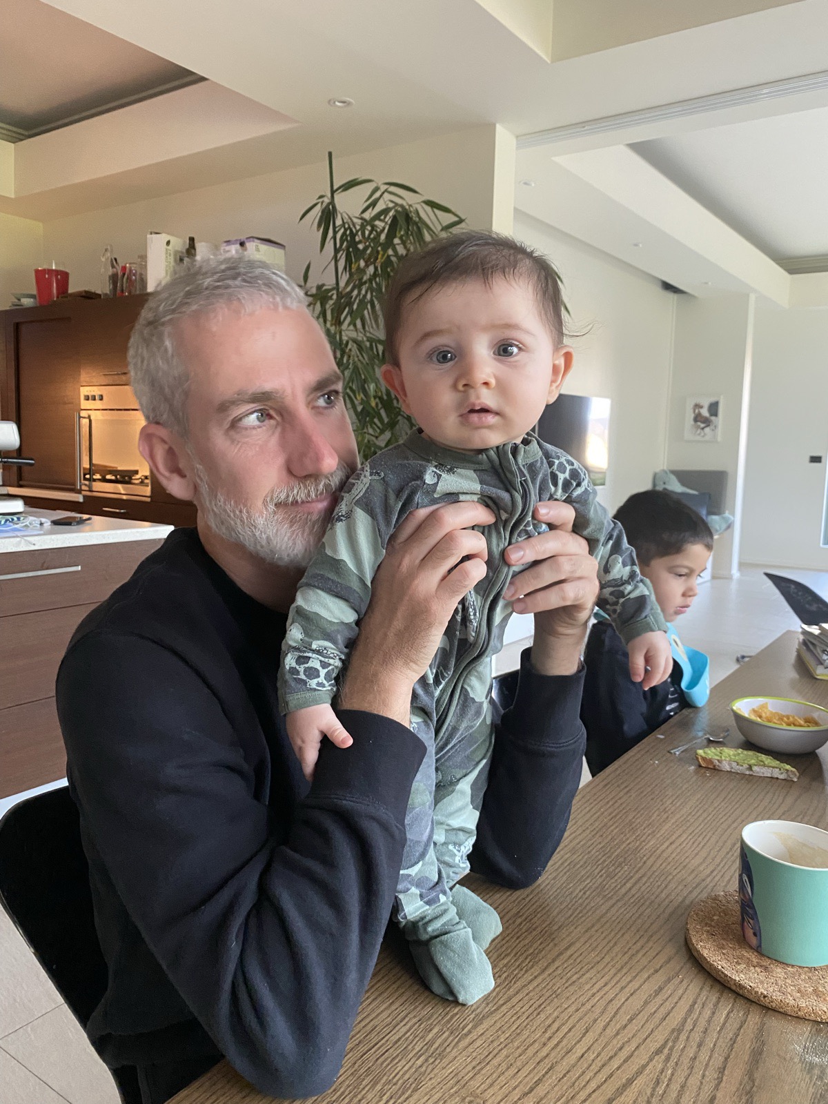 Pierre holding the baby upright at the kitchen table while his eldest has breakfast
